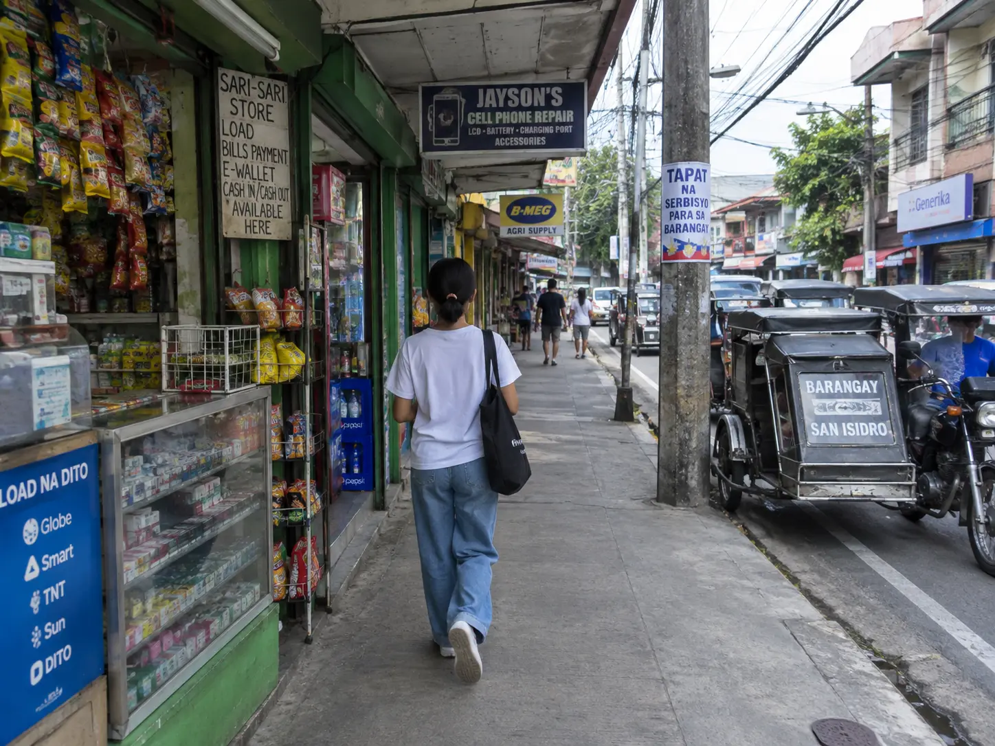 A person walking along a narrow mixed-use barangay commercial street with small shops and covered walkways